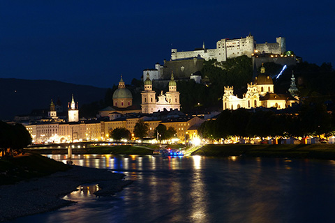A large castle overlooks a city of multiple buildings nestled behind a river at night